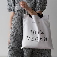 Load image into Gallery viewer, A side-view lifestyle shot of a model wearing the 100% Vegan Tote. The white canvas bag is draped over the shoulder, highlighting its spacious size and the bold "100% VEGAN" black lettering against a patterned dress.
