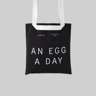 Load image into Gallery viewer, A studio shot of the An Egg a Day Tote in black canvas against a light grey background. The bag features "AN EGG A DAY" printed in bold white letters, along with the Superegg logo and "Hatched in NYC" text, showcasing a minimalist and durable carryall.