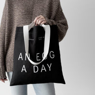 Load image into Gallery viewer, A lifestyle image of a model carrying the An Egg a Day Tote by its white handles. The black canvas bag features bold white "AN EGG A DAY" typography and the Superegg logo, paired with a cozy brown knit sweater for a minimalist and modern aesthetic.