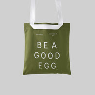Load image into Gallery viewer, A studio shot of the Be a Good Egg Tote in olive green canvas against a light grey background. The bag features "BE A GOOD EGG" printed in bold white letters, along with the Superegg logo and "Hatched in NYC" text, showcasing a durable and stylish carryall.