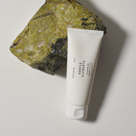 Load image into Gallery viewer, An artistic lifestyle shot of the Gentle Elements Cleansing Foam tube leaning against a textured green mineral stone. The minimalist white packaging is set against a neutral background, highlighting the cleanser’s focus on natural botanical elements and a clean aesthetic.