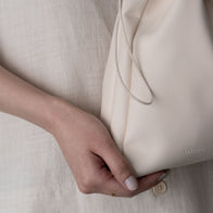 Load image into Gallery viewer, A close-up lifestyle shot of a model holding the Minimal Egg Cream Pouch by its side. The image highlights the soft, cream-colored vegan leather and the elegant drawstring cord, emphasizing its functional yet minimal aesthetic as a beauty travel essential.