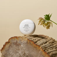 Load image into Gallery viewer, A serene lifestyle image of the Sound Renewal Serum Cream perched on a rustic wooden log. The minimalist white egg-shaped jar is accompanied by a delicate green Hellebore flower, emphasizing the product's botanical ingredients and clean beauty aesthetic.