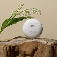 Load image into Gallery viewer, A serene lifestyle product shot of This Moment Hand Care from the Self-Care Bundle. The signature white egg-shaped container is perched on a rustic piece of natural wood, accompanied by a delicate sprig of Lily of the Valley against a warm beige background.