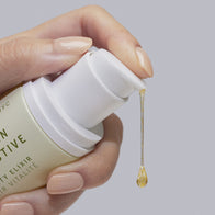 Load image into Gallery viewer, A close-up of a hand holding the Vitamin Collective Vitality Elixir bottle, with a single golden droplet of the serum falling from the pump. The image captures the viscous, nutrient-rich texture of the treatment against a soft purple-grey background.